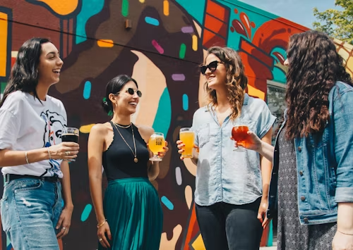 Four women with drinks in front of a colorful mural, representing vibrant outdoor social events.