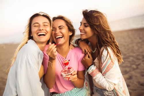 Three women laughing joyfully on a beach, representing the pure pleasure of indulgence.